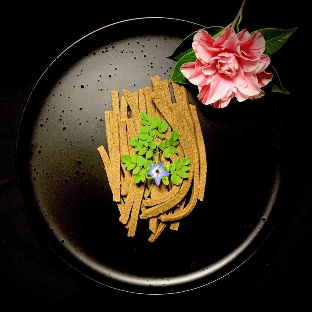 Black dish, black background, a pink candy-stripe camellia illuminates from the top right. Below, ccentred on the dish are mustard yellow, thickly cut noodles arranged neatly, except for a few artfully curved for dramatic effect. A young verdant moringa leaf adorns the noodles with a borage flower, like a blue star twinkling brightly at its base. The image is restive and serene. 

The noodles are made from ground and mushed acorns with soba flour. It is entirely gluten-free. The combination of soba (buckwheat) and acorn tastes remarkably like wheat. 