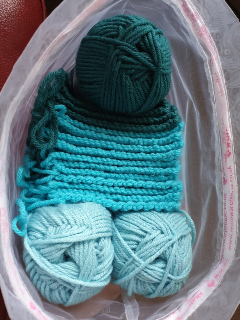 an organza bag with a gradient of sea-green wool in it. there are two light blue balls of yarn, some neatly stacked aquamarine knitted squares, and some half-knitted dark teal wool