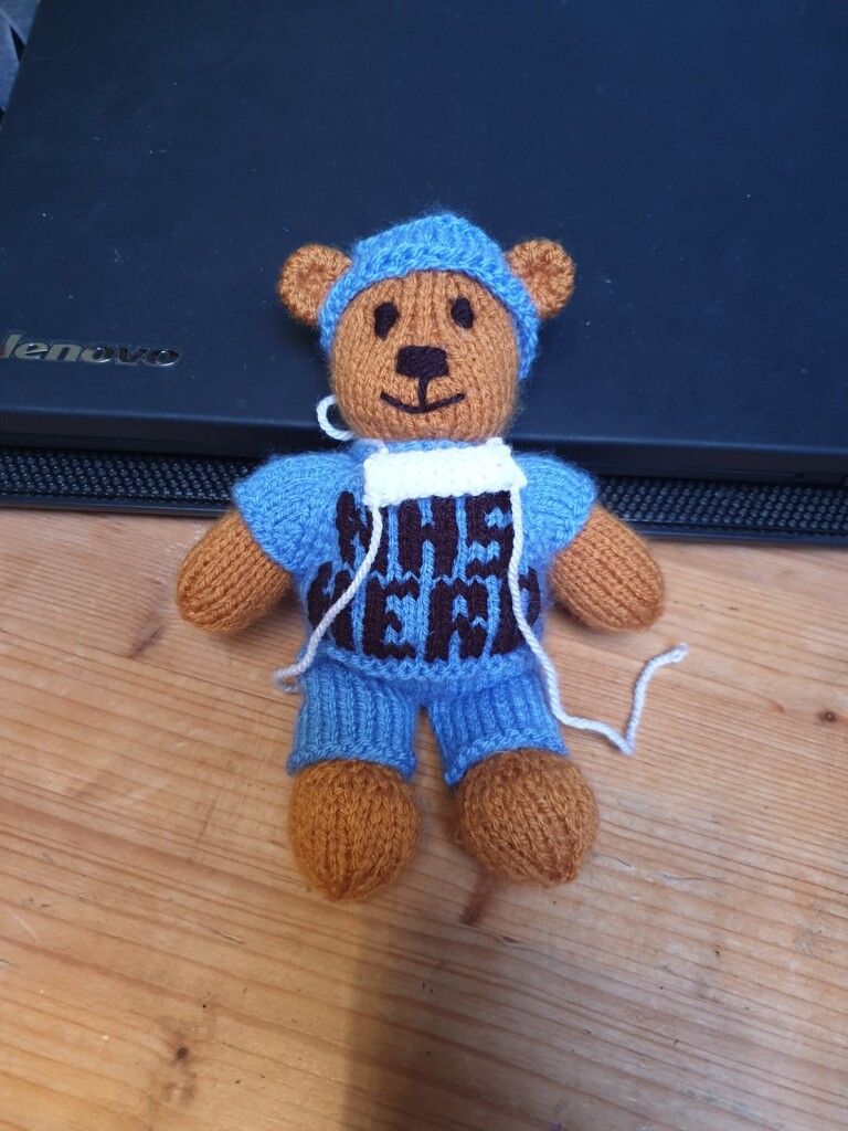 small knitted teddy with blue scrubs and NHS HERO embroidered on the front