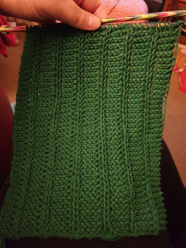 a green scarf in garter stitch with slipped purls in vertical columns, still on the needles 