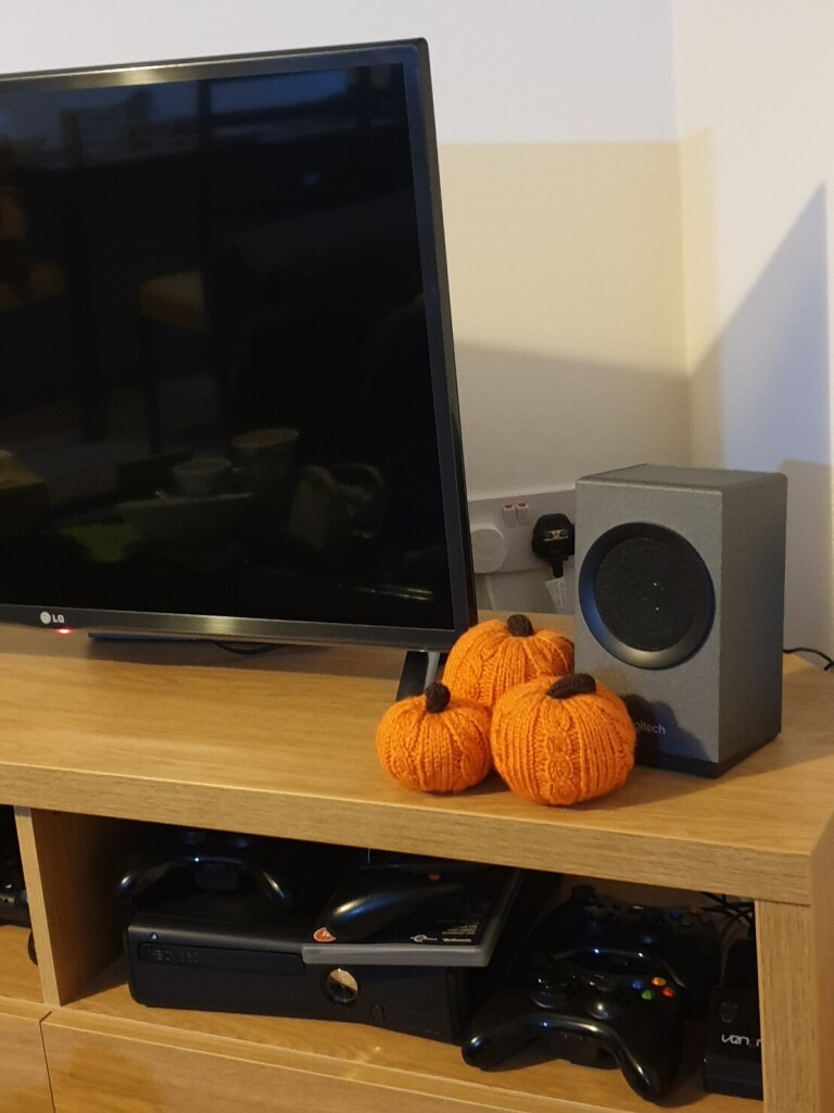 three knitted pumpkins of varying sizes sit next to a tv screen