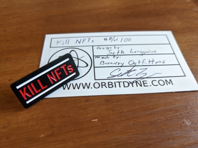An enamel pin that reads "KILL NFTS" sitting atop a business card-sized certificate of authenticity