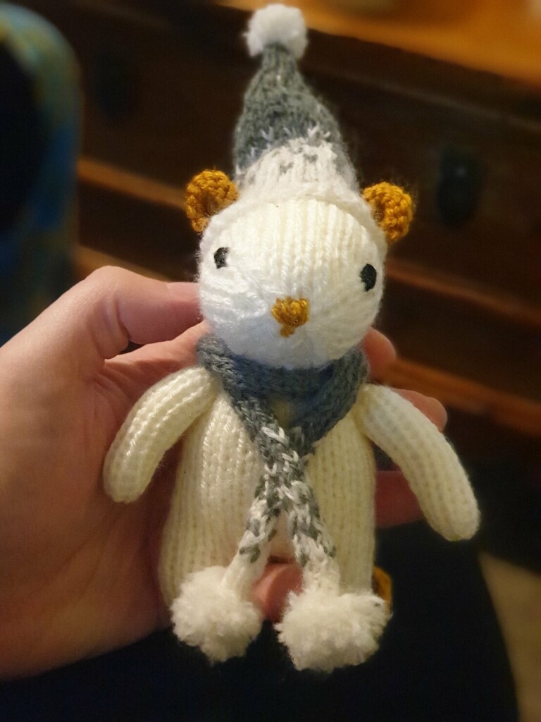 a small white knitted mouse with brown ears and nose and a little grey hat and scarf with white snow details and pompoms
