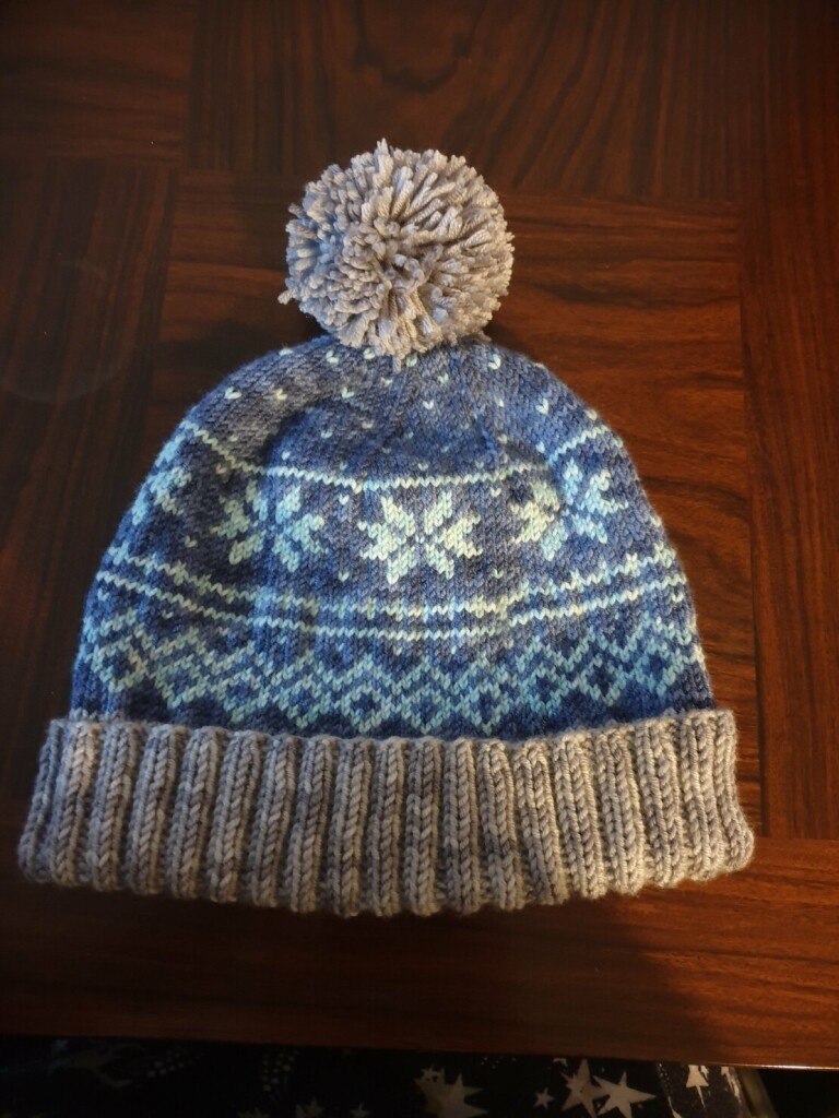 a bobble hat with grey brim and pompom and navy/light blue snowflake fairisle patterning on the body