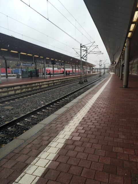 View from railway station Kassel Wilhelshöhe, Germany, platform 2, section B, heading southwards. You see the uncrowded edge of the platform and tracks, which are wet from the December snow rain. In the far background, a red regional train leaves from another platform to the south
