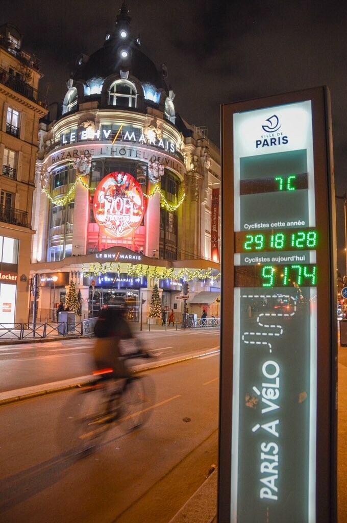 A cyclist rides on a protected bike lane past a digital counter indicating 9,174 daily and 2,918,128 annual cyclists in Paris.