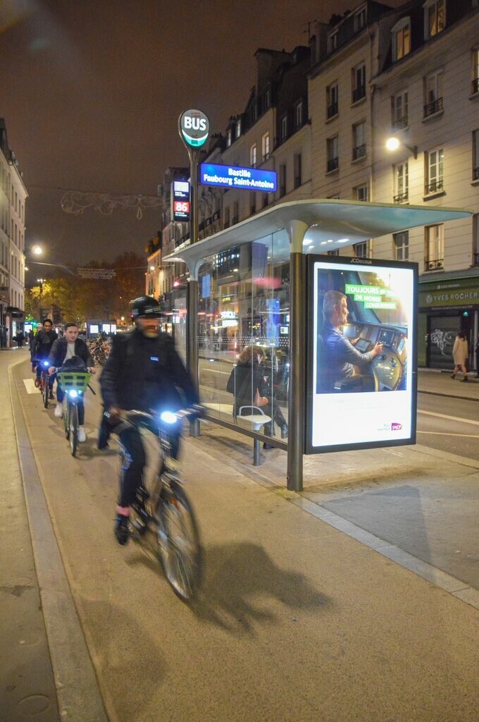 Three cyclists ride on a protected bike lane past a floating bus stop after dark on a street in Paris.