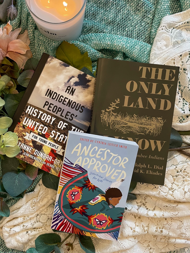 Flat lay of three paperback books on top of lace and faux florals. Included is “An Indigenous People’s’ History of the United States”, “Ancestor Approved”, and “The Only Land I Know”. 