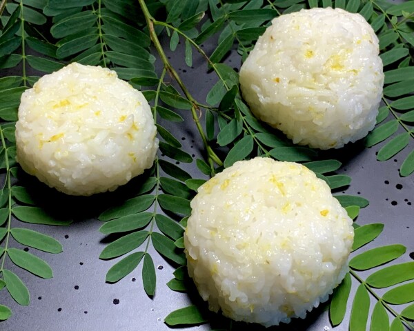 Black plate with emerald fronds of tamarind leaves cascading from the top left, three white onigiri sit, sprinkled with specks of yellow. So clean and quiescent. 