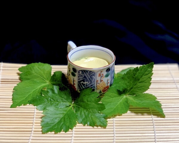 Blue background, almost black. Laying flat and forward to the viewer is a bamboo mat. Three large mitsuba leaves (like pointed clover) border a small white and blue porcelain cup. Inside the cup is a light egg custard with tiny mitsuba stems sprinkled on top. 