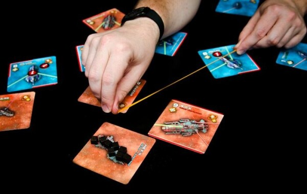 A photo of a player checking the results of combat in the game Light Speed/Stellar Conflict. There are orange and blue square cards with ships strewn about and the player is using a rubber band to continue the laser that one of the ships is firing. It is about to hit a ship with 8 black cube tokens on it.