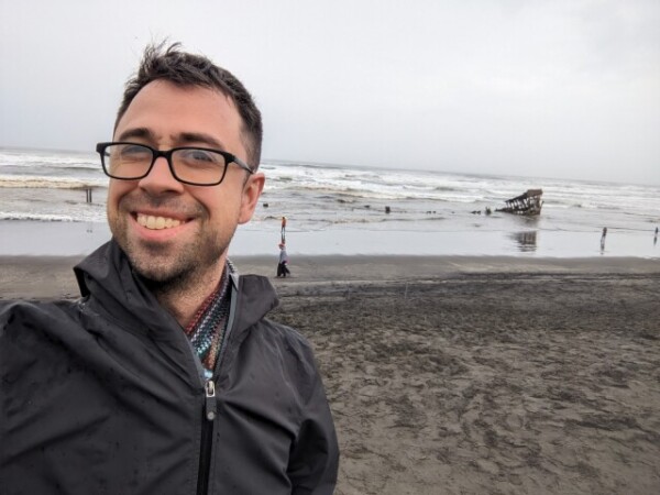 Spencer, a white masculine person with short dark hair and glasses, grins at the camera from a beach. The sky is grey and overcast and the scene seems blustery, but his smile seems to say he doesn't much mind.