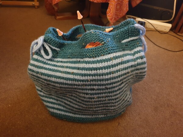 a striped, knitted bag, light blue and darker teal with light blue ties at the top. it is full of knitting
