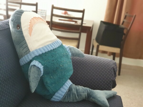 an ikea shark sitting upright on a sofa, wearing a light blue and dark teal sweater vest