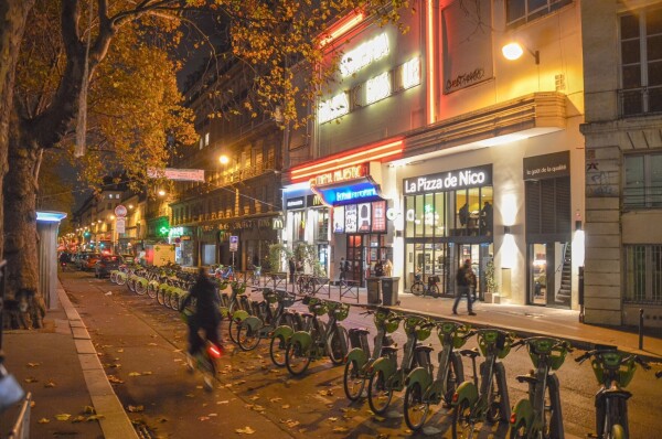 A cyclist rides along a protected bike lane lined with docked shared bikes after dark on a street in Paris.