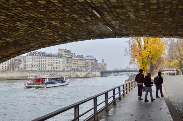 hree people walk abreast underneath a stone bridge along a car-free street on the banks of the River Seine in Paris.