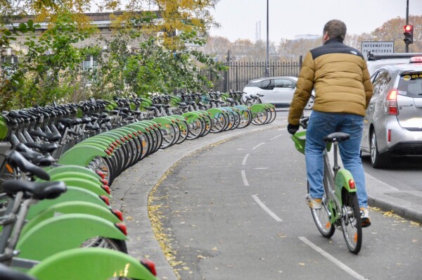 A man in winter attire rides a green and silver shared bicycle along a protected bike lane on a street in Paris.