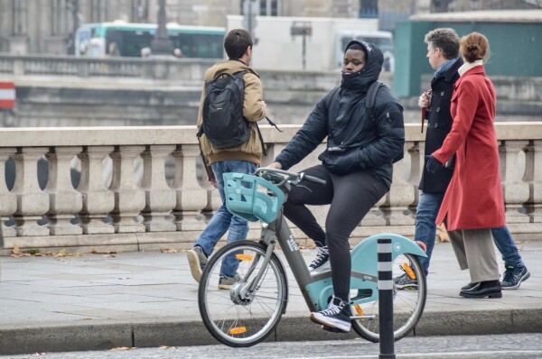 A man in winter attire rides a green and silver shared bicycle along a protected bike lane on a street in Paris.