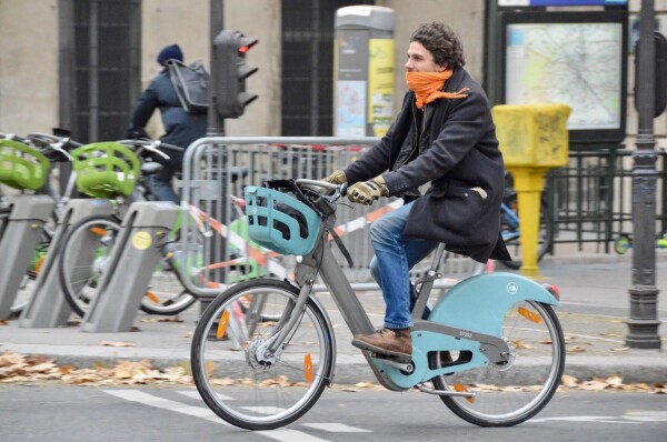 A man in winter attire rides a green and silver shared bicycle along a protected bike lane on a street in Paris.