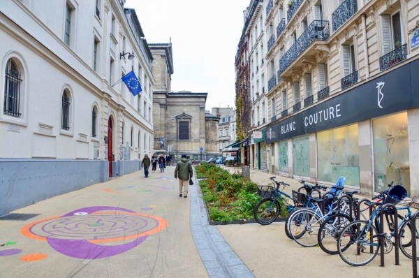 A car-free “school street” in Paris includes bike parking, vegetated areas, and white asphalt painted with colorful games.
