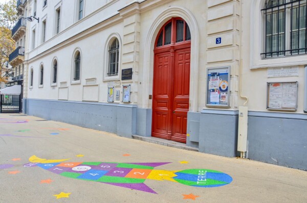 A car-free “school street” in Paris includes bike parking, vegetated areas, and white asphalt painted with colorful games.
