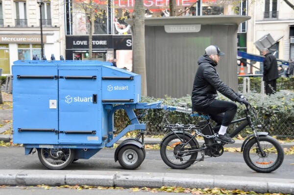 A man in casual winter attire rides an electric cargo bike on a street in Paris with a large blue trailer attached to the rear.