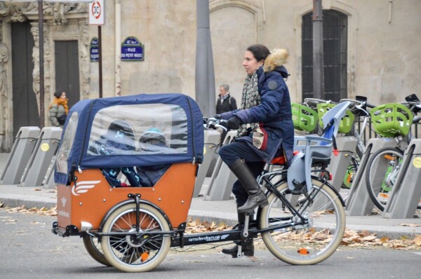 A woman in casual winter attire rides an electric cargo bike filled with small children on a street in Paris.