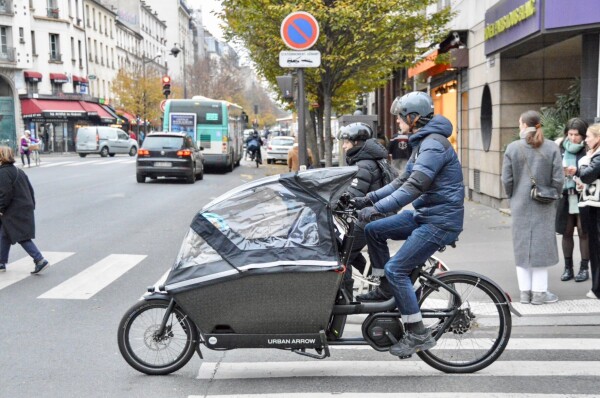 A man in casual winter attire rides an electric cargo bike filled with small children on a street in Paris.