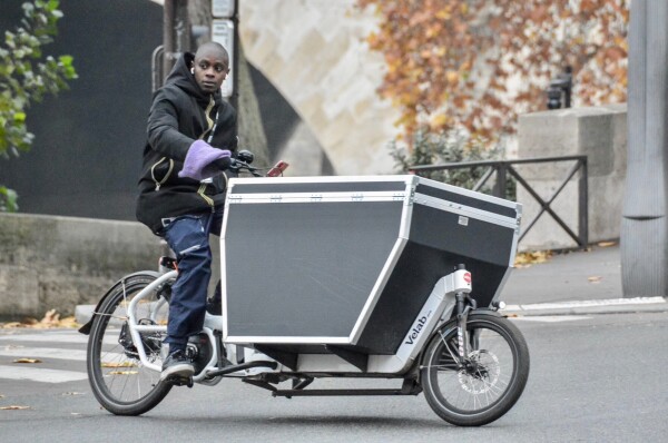 A man in casual winter attire rides an electric cargo bike carrying a large black flight case on the front on a street in Paris.