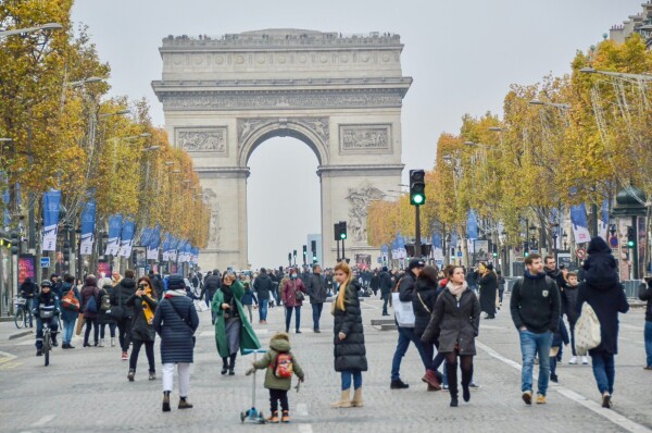 A large crowd of people walk on the car-free Avenue des Champs-Élysées in Paris. The Arc du Triomphe can be seen in the distance.