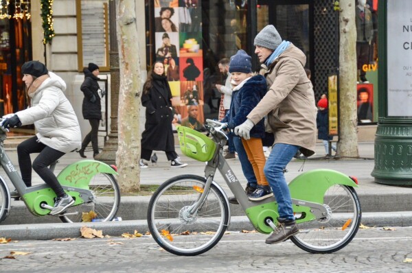 A man rides a green and silver bike-share with a child standing on the frame on the car-free Champs-Élysées in Paris.