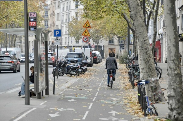 A male cyclist in casual winter attire rides on a bidirectional protected bike lane covered in fallen leaves in Paris.