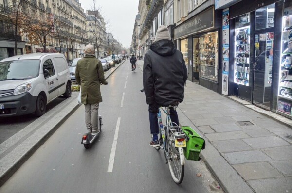 A male cyclist in casual winter attire rides on a bidirectional protected bike lane in Paris. He is being overtaken by a man on a e-scooter.