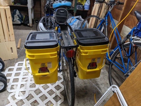 A bicycle in the garage, from the rear. Two yellow cat litter buckets, outfitted with reflectors, hang from the cargo rack.